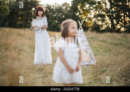 Mutter und Tochter im Sommerfeld. Konzentrieren Sie sich auf atemberaubende Boho Frau in weißem Kleid, hinter ihrer niedlichen kleinen Tochter stehen. Verwischen Sie den Blick auf ein verträumtes Mädchen, das im Gras auf dem Feld steht und wegschaut Stockfoto