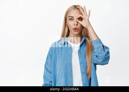 Bild der blonden jungen Frau zeigt okay Null Geste gegen das Auge, Blick durch die Finger mit beeindruckt Gesichtsausdruck, weißer Hintergrund Stockfoto