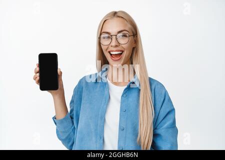 Junges blondes Mädchen in einer Brille mit Handybildschirm und lächelnd, in legerer Kleidung stehend und Smartphone-Anwendung vorführen, weiß Stockfoto