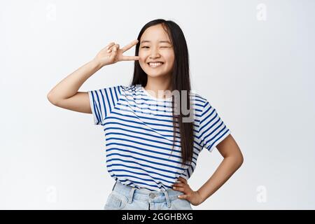 Positive und selbstbewusste asiatische Mädchen zwinkernden, lächelnd und zeigt glücklichen Frieden Zeichen in der Nähe des Auges, kawaii Pose, stehend über weißem Hintergrund Stockfoto