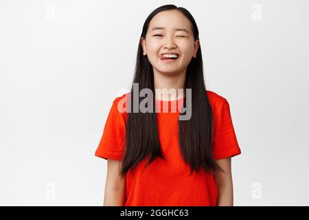 Fröhliches asiatisches Mädchen, das zwinkert und lächelt und ein glückliches Gesicht zeigt, das lässiges rotes T-Shirt trägt und vor weißem Hintergrund steht Stockfoto