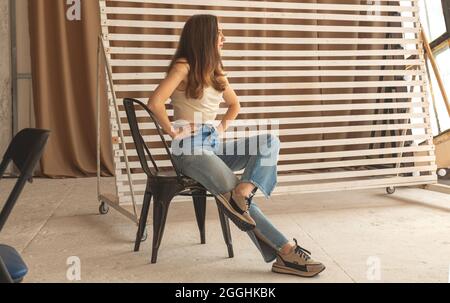 Junge Frau in legerer Kleidung posiert auf einem Stuhl im urbanen Studio-Interieur, Teenager-Mädchen in Jeans sitzt auf schwarzem Stuhl, in voller Länge Porträt-Foto Stockfoto