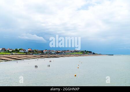 Blick auf den Kiesstrand mit Groynes und den Wohnteil der Stadt. Dunkle Regenwolken über der Stadt, mit weißem und blauem Himmel über dem Meer. Stockfoto