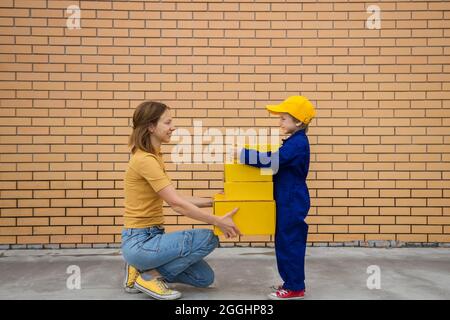 Frau und Kind, Mutter und Sohn, übergeben gelbe Kartons von Hand zu Hand. Kleiner Kurier bei der Arbeit. Express-Post, Lieferung von Glück. Mütter D Stockfoto