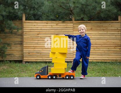 Der 5-jährige Junge in einem blauen Overall steht in der Nähe eines großen Spielzeugautos - einem Lastwagen, der mit gelben Kartons beladen ist. Kleiner Kurier bei der Arbeit. Paketzustellung, Stockfoto