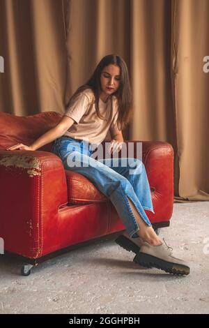 Retro-Mode. Teen Mädchen in klassischen Jeans und städtischen Sneakers sitzen auf alten Luxus-Lederstuhl im Studio. Posiert Hintergrundbild Stockfoto