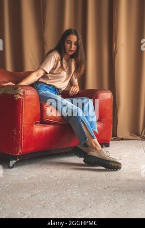 Retro-Mode. Teenager-Mädchen in klassischen Jeans und städtischen Sneakers sitzen auf alten Luxus-Lederstuhl im Studio. Posiert Hintergrundbild Stockfoto