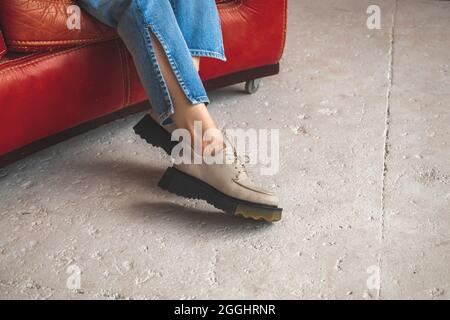 Lässige Mode mit urbanen Schuhen an den weiblichen Beinen in Denim-Jeans. Das Studio wurde auf einem roten Lederstuhl und einem Betonhintergrund gedreht Stockfoto