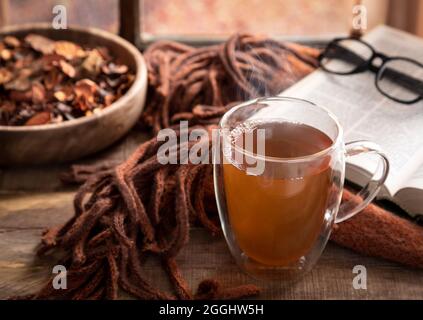 Heiße Tasse Tee mit Decke und Buch auf einem Holztisch am Fenster Stockfoto