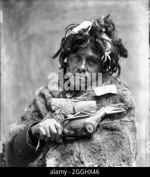 Tlingit Shaman, ca. 1900. Porträt eines Mannes, der Pelzumhang und geschnitzte Amulett-Halskette trägt, wobei er Rassel hält; Stockfoto