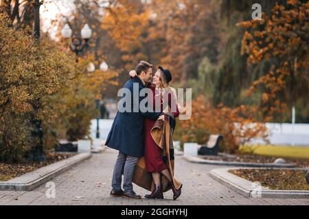 Ein junges verliebtes Paar, ein hübscher Mann und eine elegante Frau in rotem Kleid und Hutumarmungen, die an einem Herbsttag in einem Park Spaß haben Stockfoto