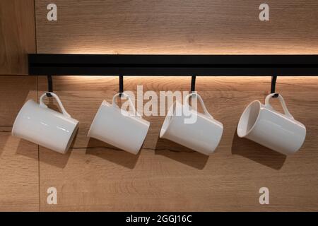 Set von leeren Trinkbecher aus weißer Keramik, die auf einem Ständer in der Küche hängen, auf einem Holzhintergrund mit Licht. Stockfoto