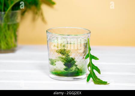 Grünes Chlorophyll-Getränk im Glas mit Wasser auf weißem Tisch, orangefarbener Hintergrund. Antioxidative Lebensmittel und Getränke, Detox-Diät Stockfoto