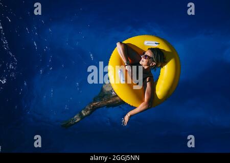 66-jährige Frau genießt einen sonnigen Tag im Pool mit einem gelben Schwimmer Stockfoto