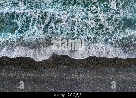 Stürmische grüne Wellen mit weißem Meeresschaum, der am dunklen Sandstrand bricht, Blick von direkt oben Stockfoto