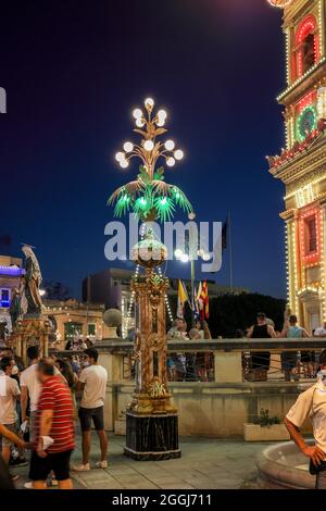 MOSTA, MALTA - 15. Aug 2021: Pilaster unterstützt verschiedene farbige Lichter als Straßendekoration. Menschen in der Umgebung. Anlass des Titularfestes von Mosta, t Stockfoto