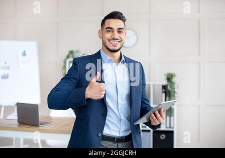 Lächelnder arabischer Geschäftsmann, der den Computer hält und im Büro mit dem Daumen nach oben zeigt Stockfoto