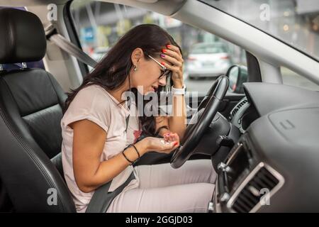 Frau in Dispair hält ihren Kopf auf dem Fahrersitz sitzend, mit einer Hand voll Pillen in ihrer anderen Hand. Stockfoto