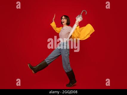 Herbstwetter. Junge Frau in Regenmantel und Gummistiefel kämpfen Wind und gehen über roten Hintergrund Stockfoto