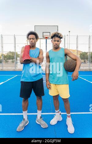 Coole afroamerikanische Männer halten den Basketballball lächelnd und schauen auf die Kamera im Basketballfeld. Stockfoto