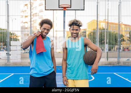 Coole afroamerikanische Männer halten den Basketballball lächelnd und schauen auf die Kamera im Basketballfeld. Stockfoto