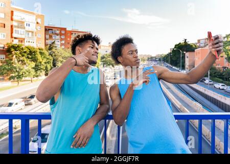 Zwei junge afro-amerikanische Männer mit blauem ärmellosem T-Shirt, die ein Selfie auf der Fußgängerbrücke mit dem Highway unten in der Stadt machen. Stockfoto