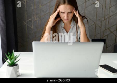 Depressive junge Geschäftsfrau, die den Kopf in den Händen hält, hat Probleme und starrt verzweifelt auf den Laptop-Bildschirm. Eine Frau im formellen Hemd hat bei einer Arbeit einen Fehler gemacht Stockfoto
