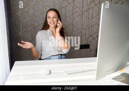 Glückliche weibliche Büroangestellte chattet auf dem Smartphone, das am Schreibtisch vor dem Laptop im Heimbüro sitzt. Fröhliche Frau im lässigen Hemd hat angenehme Telefongespräche, lacht und gestikiert Stockfoto