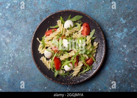 Draufsicht auf leckere vegetarische Pasta mit Kirschtomaten, Weichkäse-Mozzarella und grünen Rucola-Blättern auf blauem Beton-Hintergrund Stockfoto