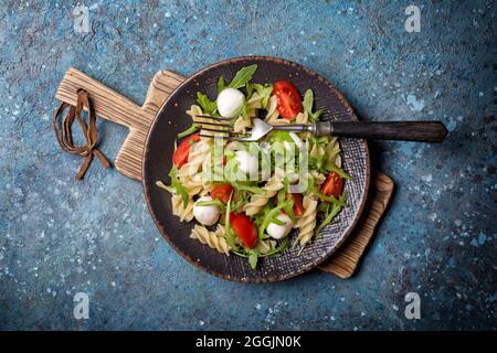 Draufsicht auf leckere vegetarische Pasta mit Kirschtomaten, Weichkäse-Mozzarella und grünen Rucola-Blättern auf blauem Beton-Hintergrund Stockfoto