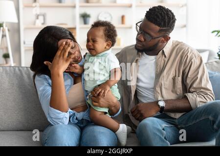 Müde afroamerikanische Eltern sitzen mit weinenden Kind auf dem Sofa Stockfoto