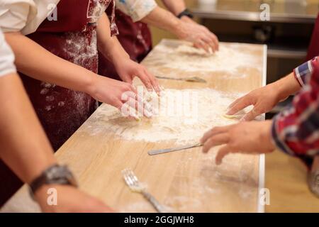Die Hände mehrerer Personen kneten den Teig auf dem Tisch. Ein rohes Ei mit Mehl vermischen. Nahaufnahme. Stockfoto