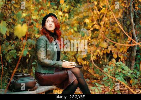 Nette Frau mit grafischem Tablet auf Parkbank. Herbstporträt. Rothaarige Mädchen in Baskenmütze und modische Jacke schaut nach oben. Designer oder Freiberufler. Lederrucksack auf Holzbank. Stockfoto