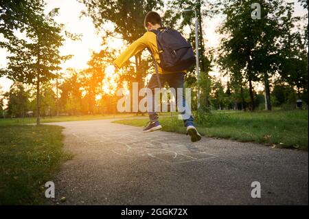 Ganzkörperportrait eines aktiven Schuljungen, der nach dem ersten Schultag seine Freizeit genießt und auf dem Boden hoppscotch spielt. Straßenspiele für Kinder Stockfoto