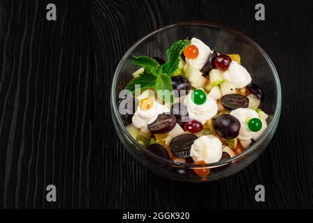 Salat aus Trauben Äpfel Birnen Kiwi Orangen mit Mascarpone Chease und Creme. Gesunder, frischer Obstsalat im Glas auf schwarzem Holzboden Stockfoto