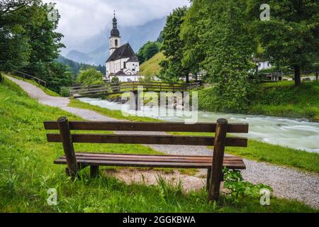 Eine Holzbank mit Pfarrkirche, Fluss abd Berge im Hintergrund. berchtesgaden, Ramsau Deutschland Stockfoto