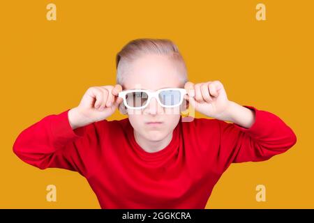 Ein Kind in einer roten Jacke hält seine Hände hinter seiner Brille auf einem gelben Hintergrund Stockfoto