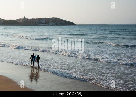 Von ihrem Rücken aus gesehen, wie das junge Paar das Wasser betrat, um bei Sonnenuntergang am Strand von Gijon zu surfen. Blick auf die Stadt in der Ferne. Asturien, Spanien, Europa Stockfoto