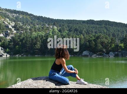 Schwarze Frau mit lockigen afro Haaren sitzt auf einem Felsen. Beobachtung der Schwarzen Lagune und des Pinienwaldes in Soria, Spanien. Stockfoto