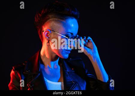Nachtportrait der Frau. Neonlicht. Positive Einstellung. Selbstbewusste glückliche Frau, in Lederjacke und Sonnenbrille, lächelnd in rot-blauem Glanz isoliert auf schwarzem Hintergrund Stockfoto