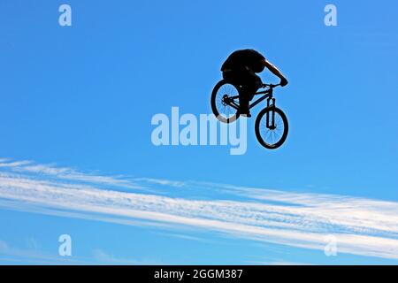 Silhouette von BMX oder Mountain Biker hoch in der Luft während einer Stunt-Show, die vor einem hauptsächlich hellblauen Himmel mit einem Band weißer Wolken spielt Stockfoto