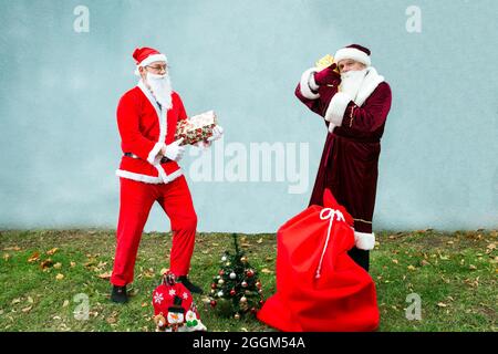 Zwei Weihnachtsmann sammeln einen großen Beutel mit Geschenken für Weihnachten. Nahaufnahme auf grauem Hintergrund und grünem Gras. Stockfoto