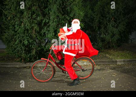 Der Weihnachtsmann fährt mit dem Fahrrad und trägt eine große Geschenktüte. Nahaufnahme auf dem Hintergrund von Nadelbäumen. Stockfoto