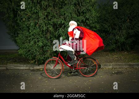Der Weihnachtsmann fährt mit dem Fahrrad und trägt eine große Geschenktüte. Nahaufnahme auf dem Hintergrund von Nadelbäumen. Stockfoto