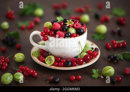 Beerenmischung in einem Becher. Kirschen, Himbeeren, Stachelbeeren und rote Johannisbeeren auf braunem Hintergrund. Berry Stillleben. Sommerkomposition. Stockfoto