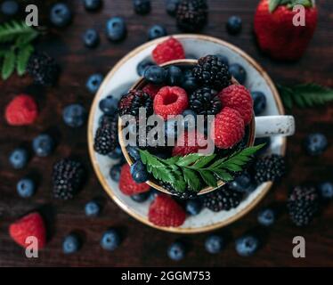 Beerenmischung in einer Tasse auf dunklem Grund. Himbeeren, Brombeeren und Heidelbeeren in einem Becher in der Nähe. Schöne Sommer-Beere Zusammensetzung. Draufsicht, FLA Stockfoto