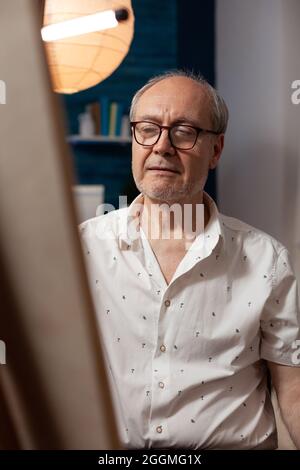 Portrait eines älteren Künstlers, der die Leinwand mit einem Meisterwerk betrachtet, das zu Hause in der Werkstatt sitzt. Nahaufnahme eines älteren Mannes mit künstlerischem Hobby, der über das Zeichnen mit professionellen Instrumenten nachdenkt Stockfoto