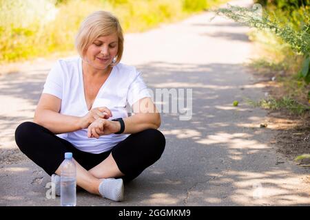 Das Foto einer fröhlichen, blonden, sportlichen Frau, die in einer Lotusposition auf der Straße im Park sitzt und Sportkleidung trägt, die auf die Digitaluhr zeigt Stockfoto