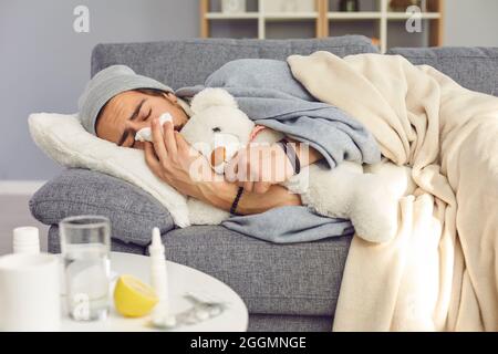 Kranker junger Mann, der zu Hause auf der Couch liegt, Teddy umarmt und Nase in Papiertuch bläst Stockfoto