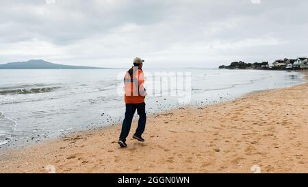 Mann, der bei Sonnenaufgang am Strand von Milford mit Rangitoto Island in der Ferne, Auckland, läuft Stockfoto